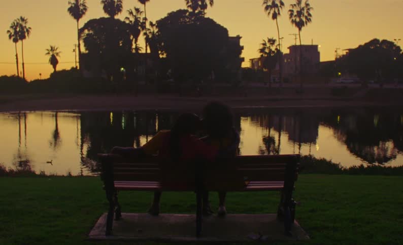 Couple sit together on park bench looking at pond that reflects red sky and nearby trees