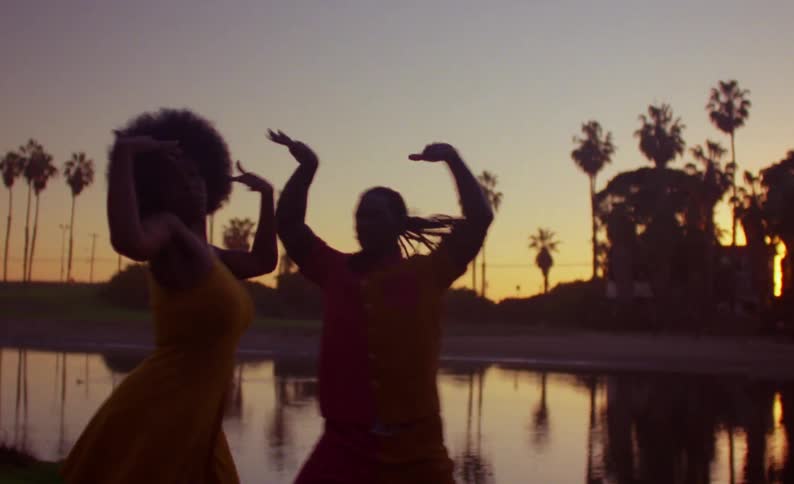 Female dancer with yellow dress and curly hair and male dancer with braids and red t shirt dance on lake bank at dusk