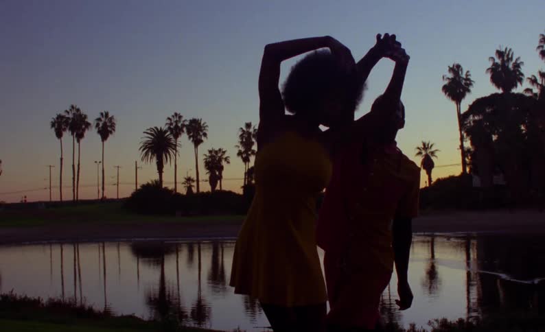 Male and female dancers dance in park on shore of pond under a red tinted sky