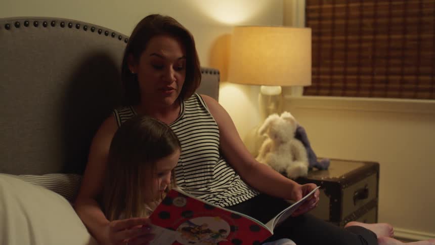 A woman reads a book to a little girl as they sit together on a bed in a bedroom