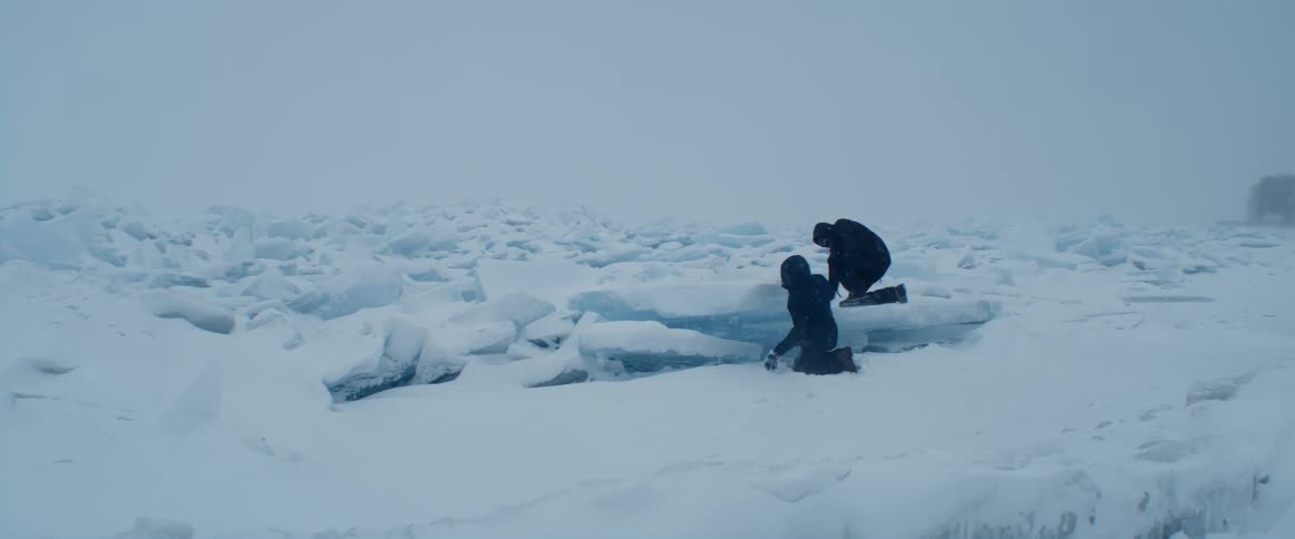 Two people wearing black clothes are in the arctic snow and one person is looking under a piece of ice as the other stands on pile of broken ice