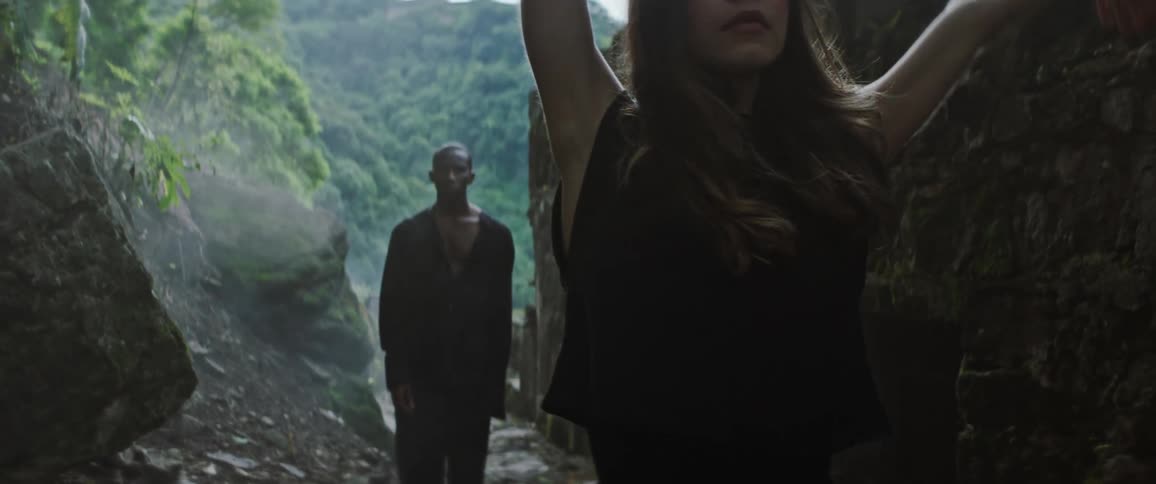 A young woman dances next to a stone fort at the base of a cliff as a man stands behind her watching