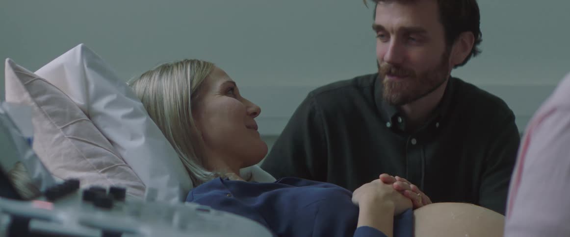A woman lies with pregnant belly exposed in an exam room holding hands with and smiling at her husband