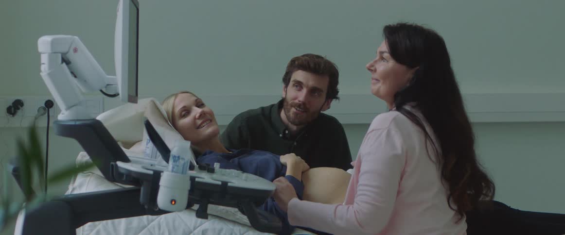 A man sits next to his wife as she lies in a bed while a woman runs an ultrasound probe over her pregnant stomach