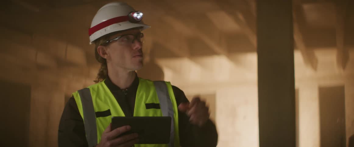Construction worker wearing bright yellow vest and white hardhat supervising a construction site at night