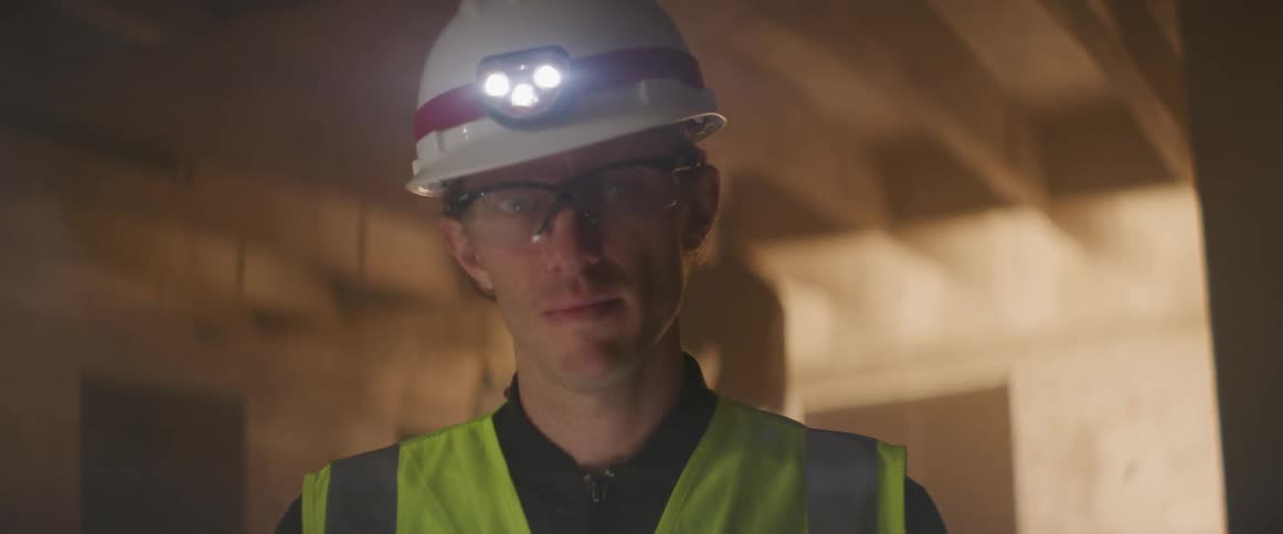 A worker on a construction site adjusts his hardhat before turning to look into the camera