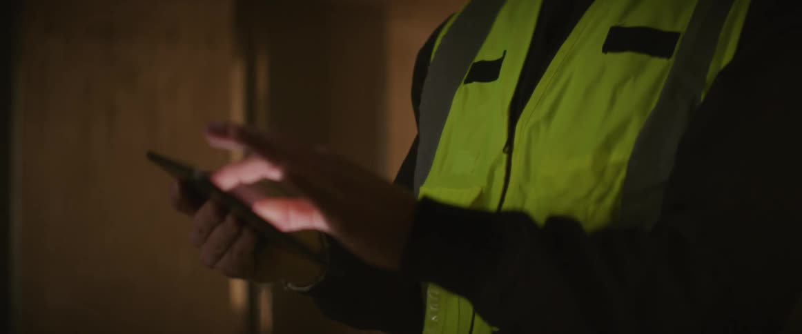 A man wearing a hardhat with light and safety vest holds up his phone to take a photo