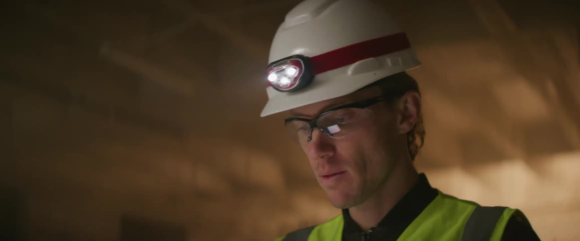 Construction worker wearing bright yellow vest and white hardhat supervising a construction site at night with a tablet