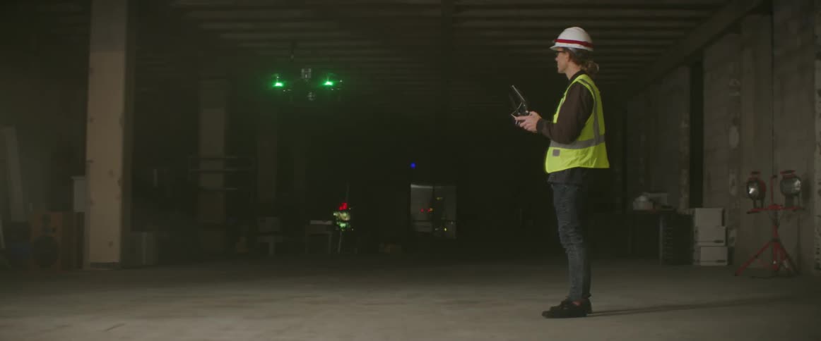 A man wearing a hardhat and safety vest controls a drone flying with green light glowing