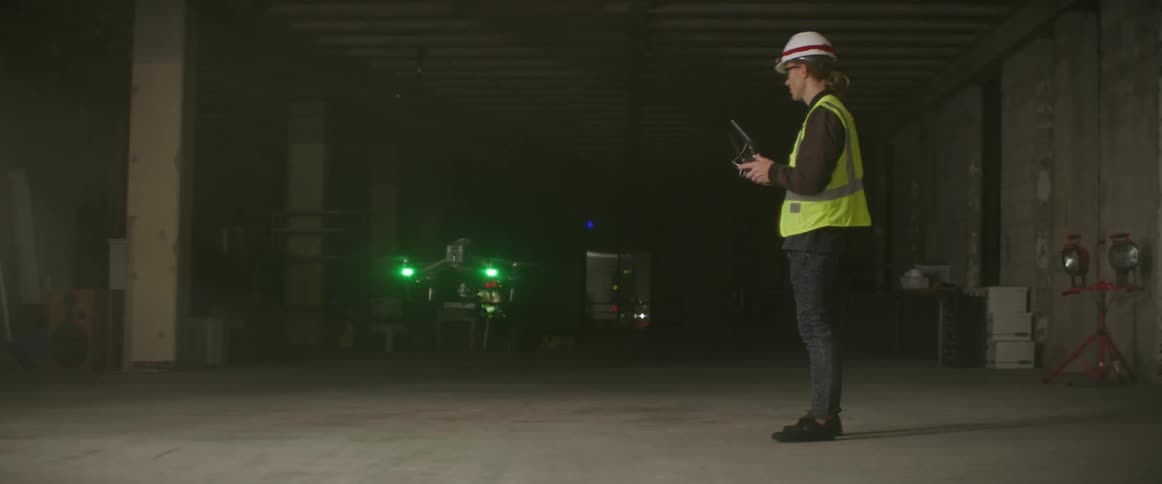 Worker wearing safety vest stands in building and manipulates drone with controller