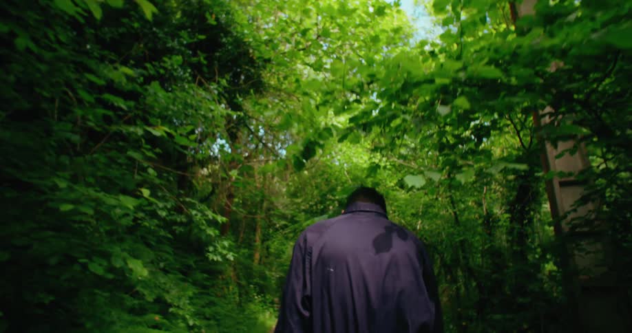 A man walks slowly into the trees passing a tall post with a cross