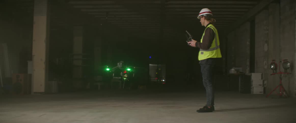 A drone with green lights glowing flies past a man holding the remote