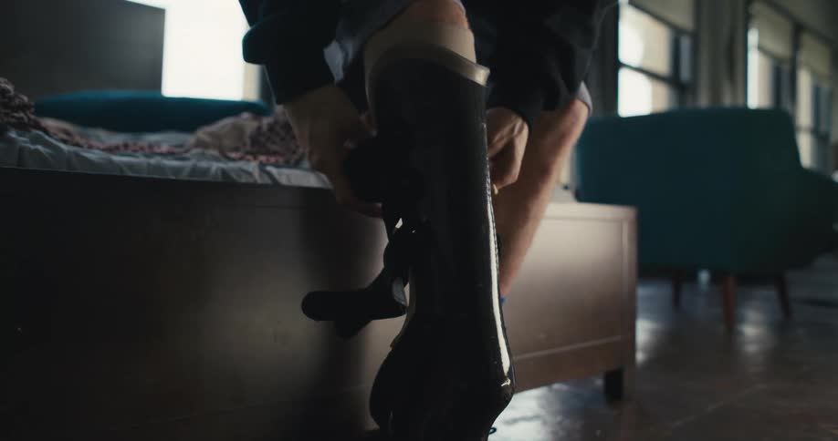 A young man sits on his bed at home and then stands up and straps on his artificial leg and walks away