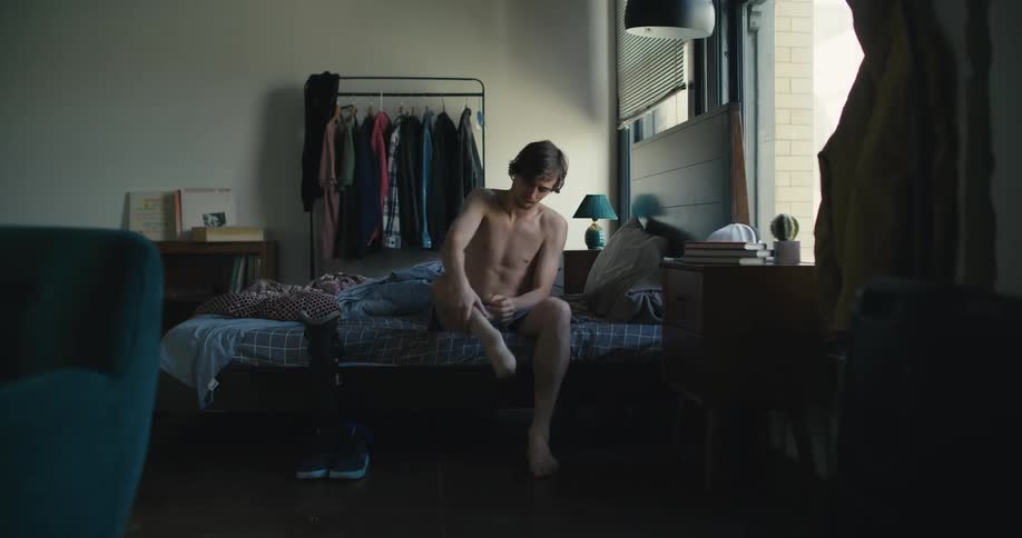 Man sits on side of bed after checking time and puts on his prosthetic