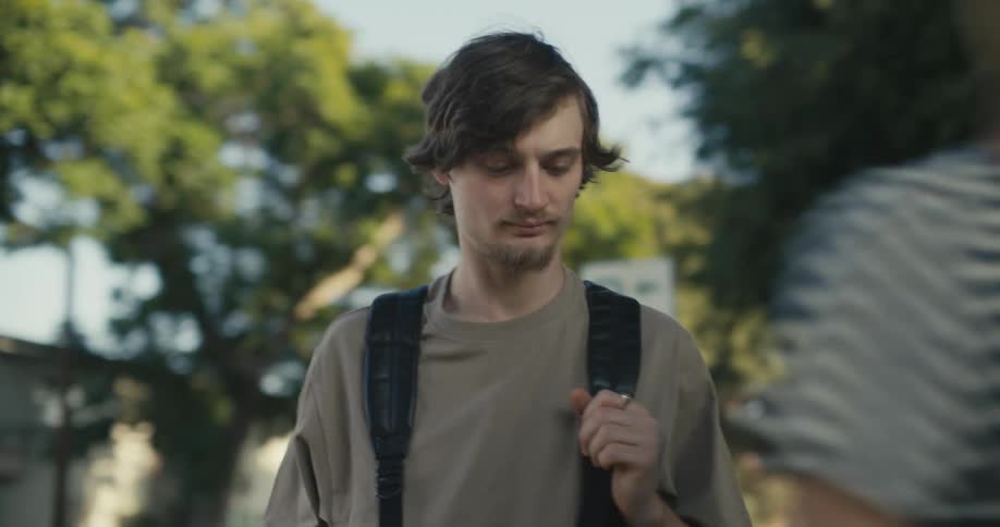 A teenager is standing outside holding the strap of his backpack and looking around as two people pass by him