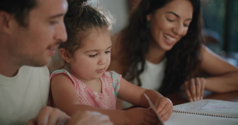A man and woman sit at a table at home with their young daughter as she flips through a notebook
