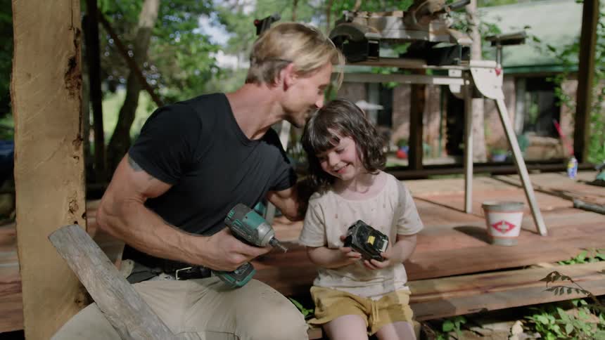 A man shows his daughter how to attach a battery to a drill and kisses her on the forehead