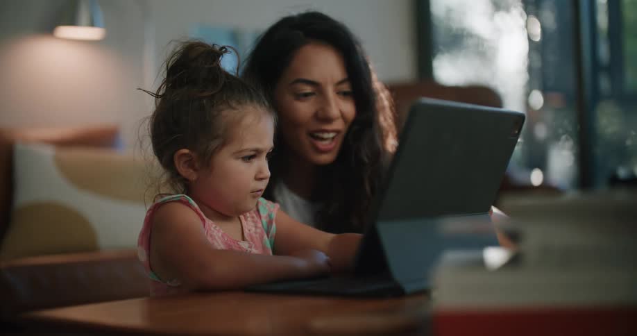 Mother with wavy hair helps daughter use tablet sitting at coffee table in living room at home