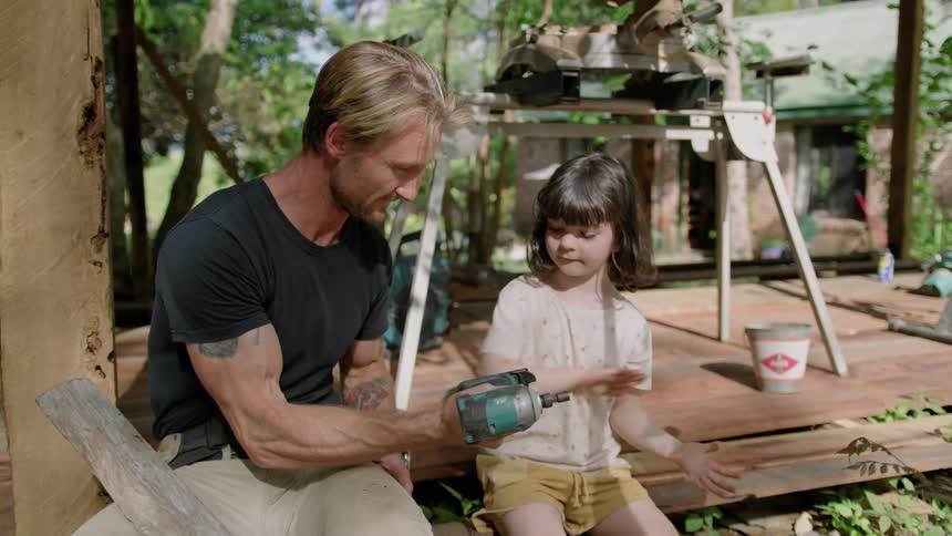 A girl smacks a drill battery into place for her dad as they sit outside