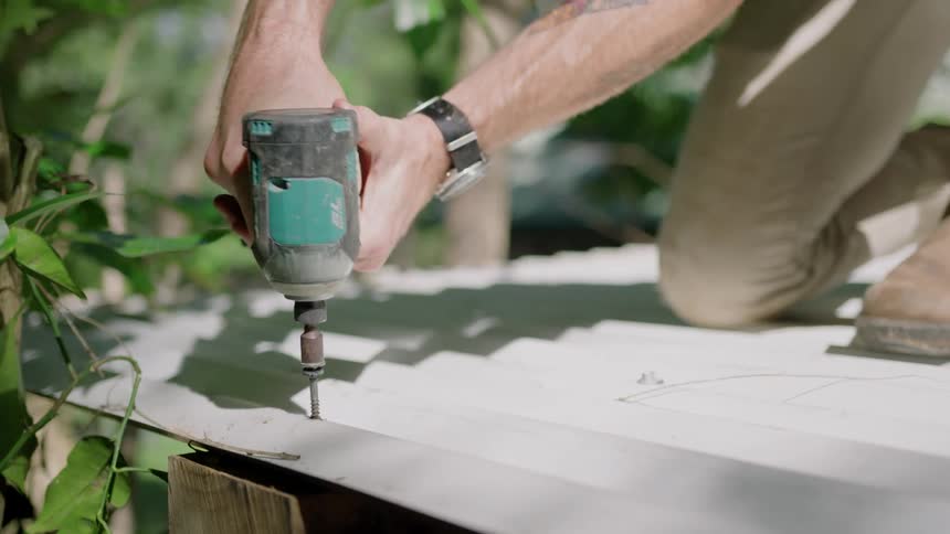 A man uses a power drill to secure the metal roof on a shed