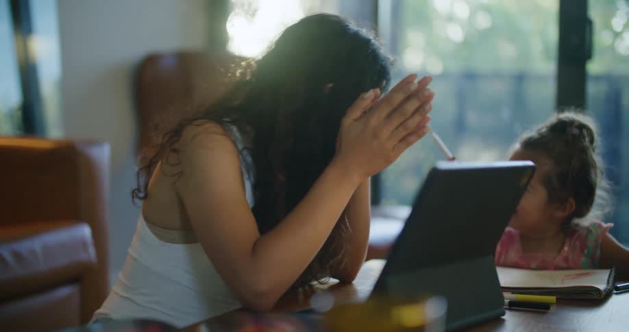 Mother with wavy hair and sleevelesss white shirt uses tablet sitting at coffee table next to daughter making a drawing in notebook in living room