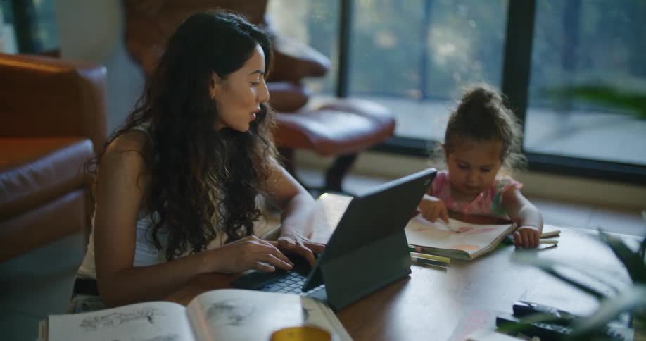 Mom with wavy hair and white shirt uses tablet and helps daughter make a drawing in notebook at coffee table in living room at home