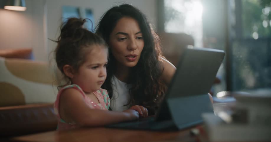 A woman sits on the floor with her daughter as the girl uses a tablet
