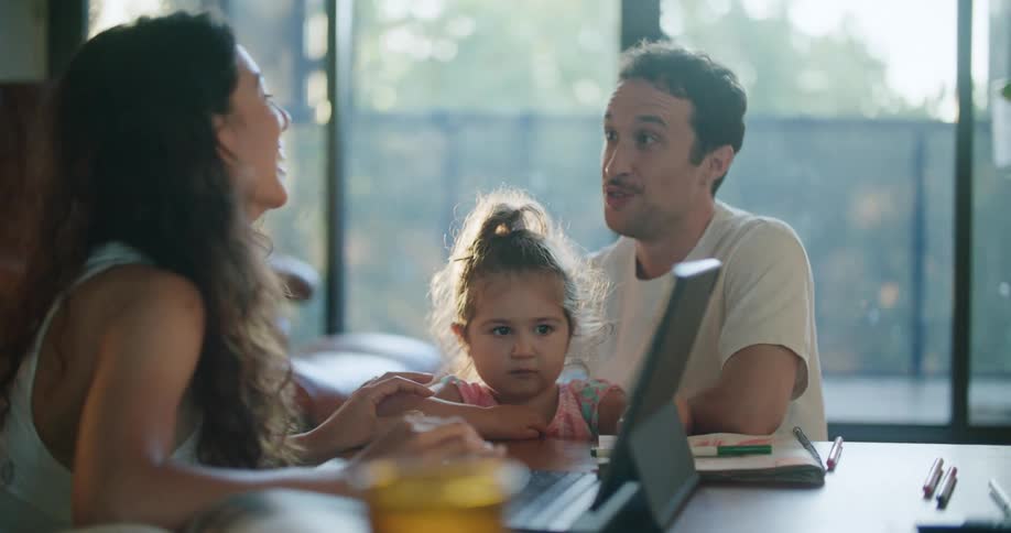Father with mustache white t shirt holding toddler girl talks with mother with wavy hair using tablet at coffee table