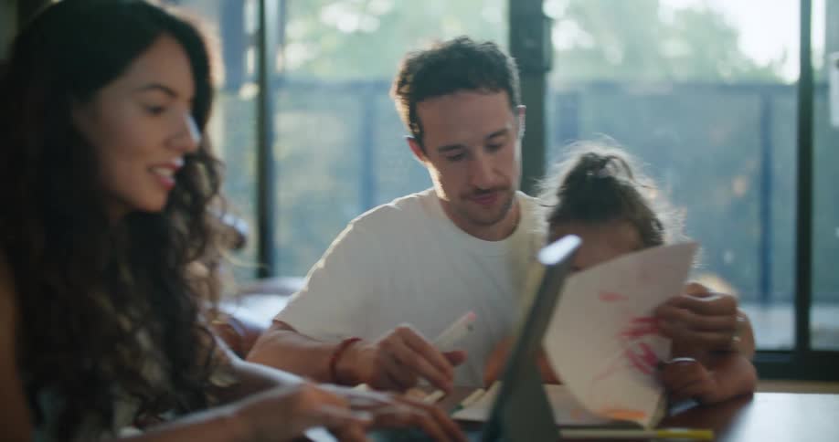 Father with mustache white t shirt holding toddler girl making a drawing in notebook next to mother with wavy hair using tablet at coffee table