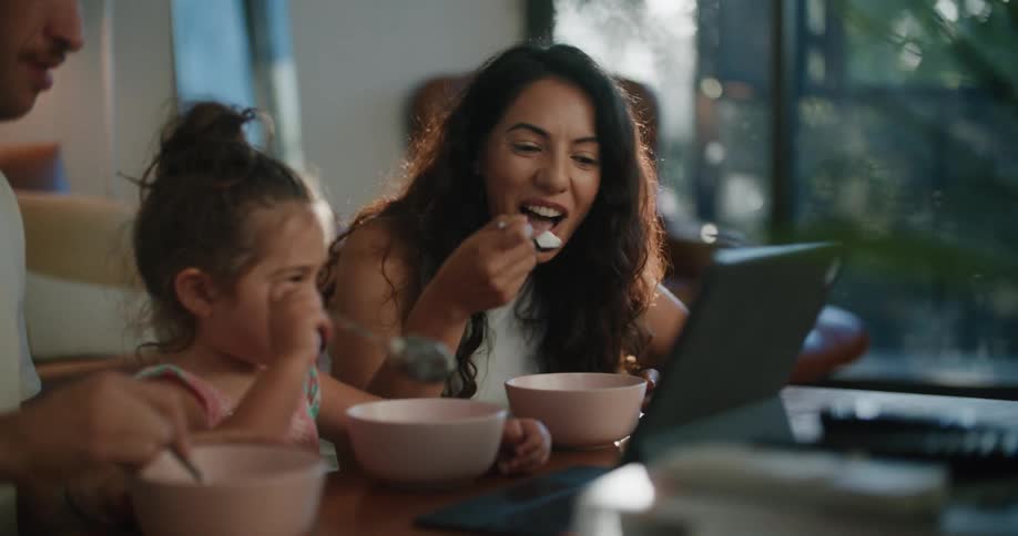 A family sit together eating from bowls while watching a tablet screen