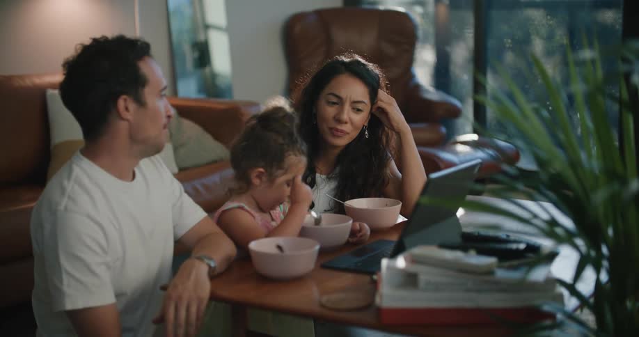 A couple chat while their daughter watches a laptop as they sit and eat on the floor