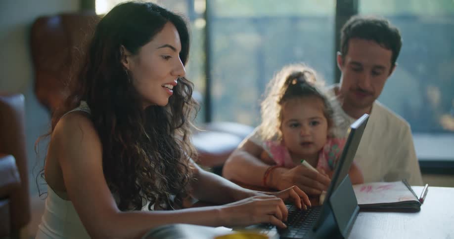 Father with mustache white t shirt holding toddler girl making a drawing in notebook next to mother with wavy hair using tablet at coffee table