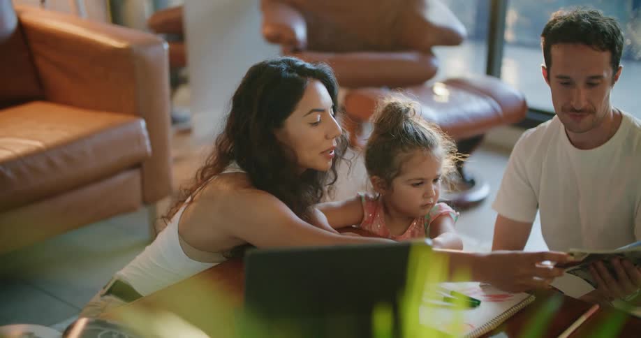 Toddler girl leans against coffee table between father with mustache white t shirt and mother with wavy hair and sleeveless white shirt reading magazine in living room at home