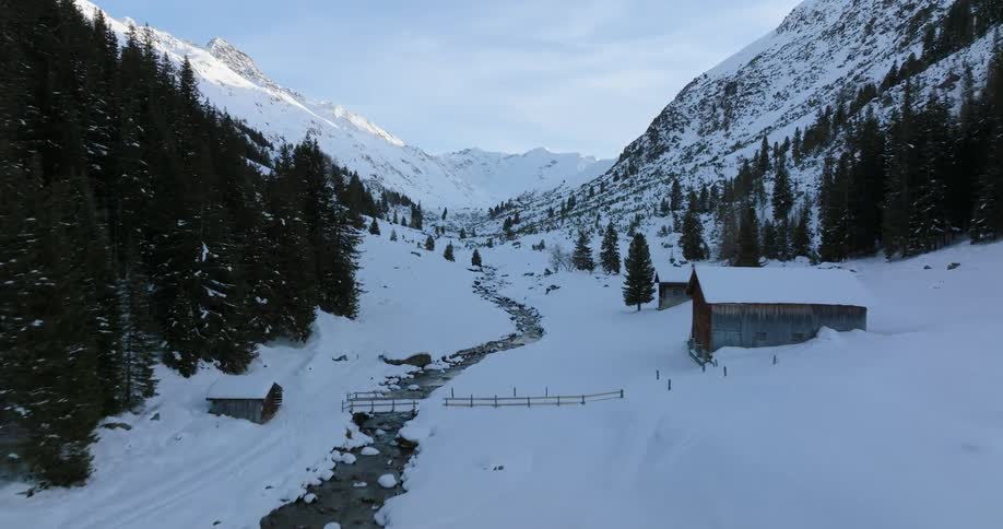 A mountain stream winds through a valley past a cabin shelter with snow covered bridge