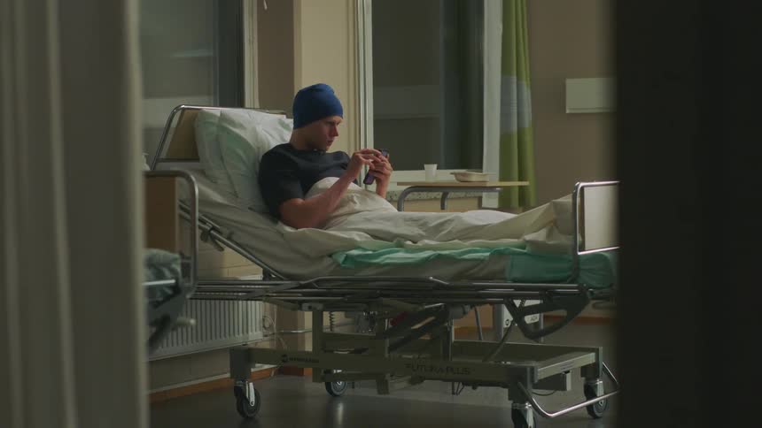 A man sitting in a hospital bed reaches down and gets his cell phone and smiles and waves as he answers it to video chat