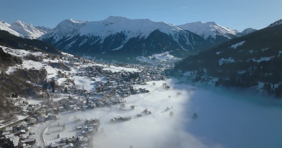 A layer of fog hangs over a snow covered valley village with mountains surrounding