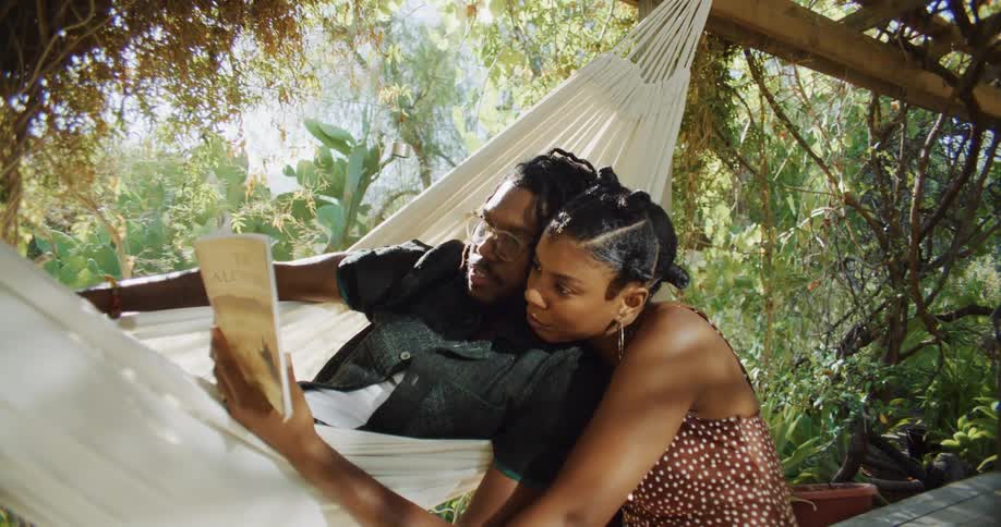Woman sits next to man lying in a hammock as they read a book together