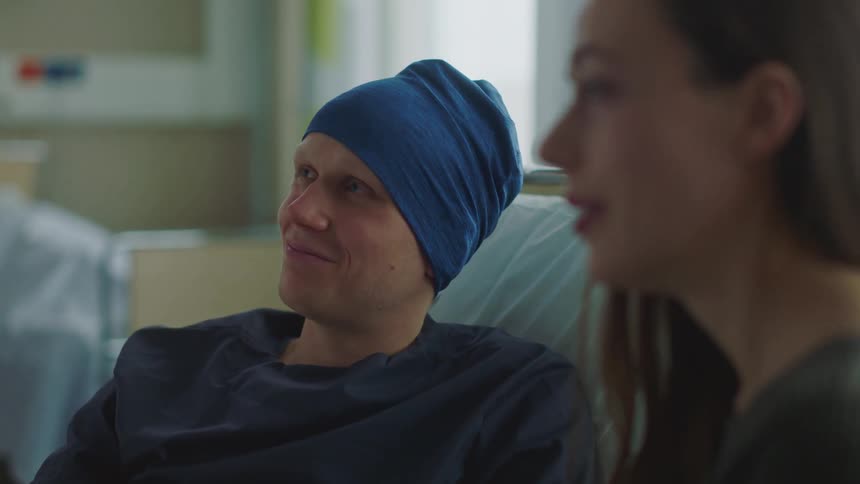 A woman sits next to a man in a hospital bed and they nod their heads as they listen to someone and then look at one another and smile and hug