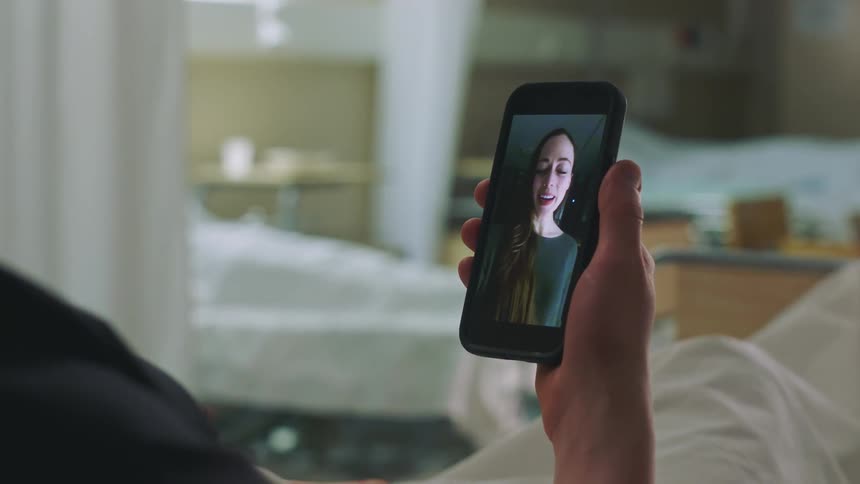 A man lying in a hospital bed holds his cell phone and waves as he video chats with a woman