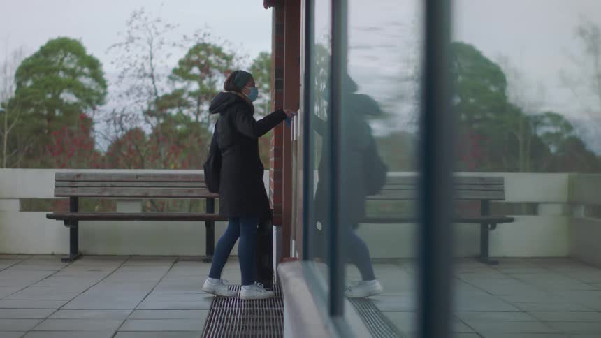A woman wearing a coat and a face mask walks up to a door and swipes her key card and then goes inside