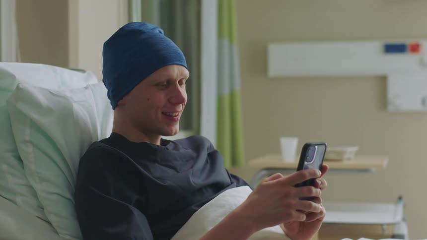 A man is sitting up in a hospital bed smiling as he talks on his phone