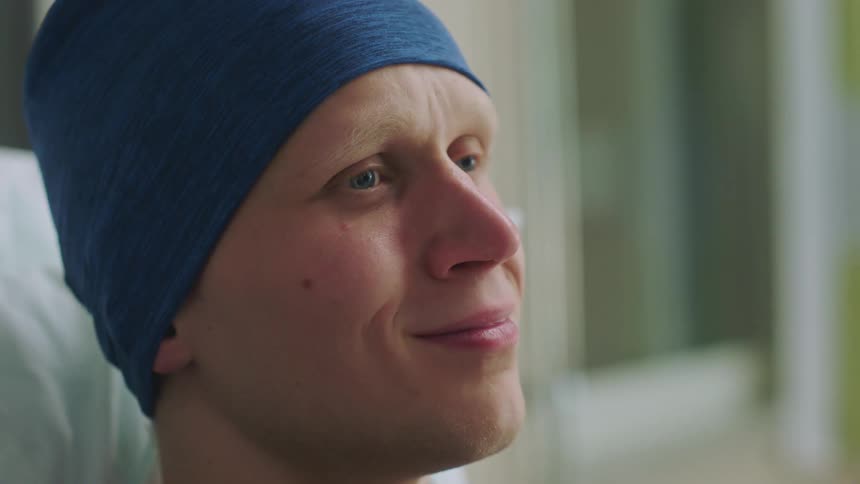 Patient holding back tears wearing a blue cap over his head resting on a bed in a hospital