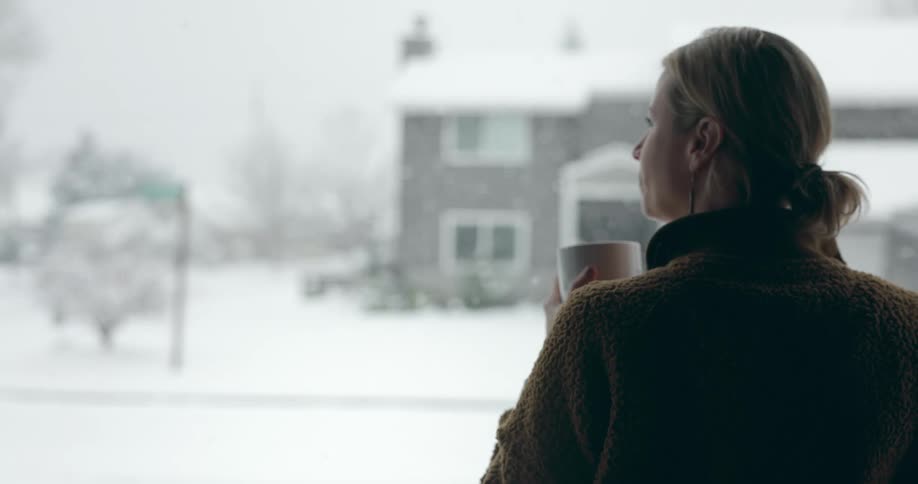 Blonde woman with brown jacket drinks from cup and looks out window while snowing