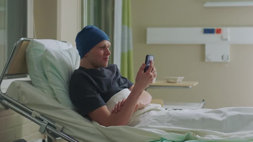 Young man laying in a hospital bed picks up a smartphone to answer a call