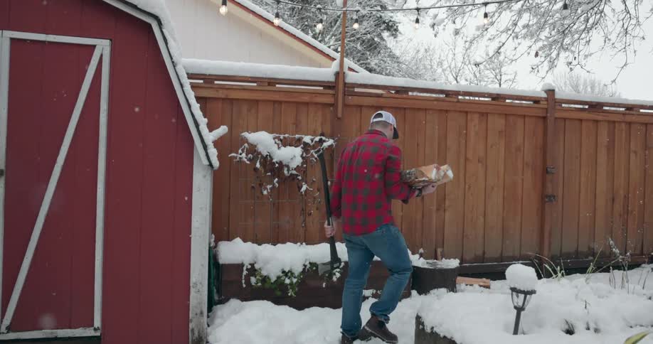 A man carries wood in his back yard while snow falls