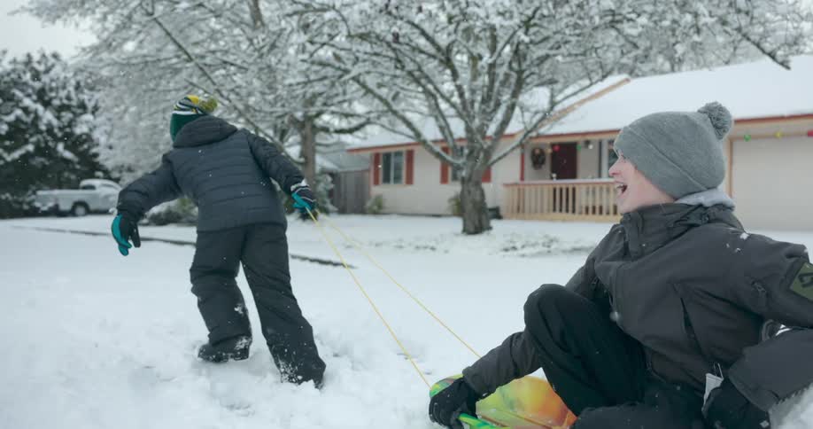 A boy is pulling another boy on a sled through the snow in the street