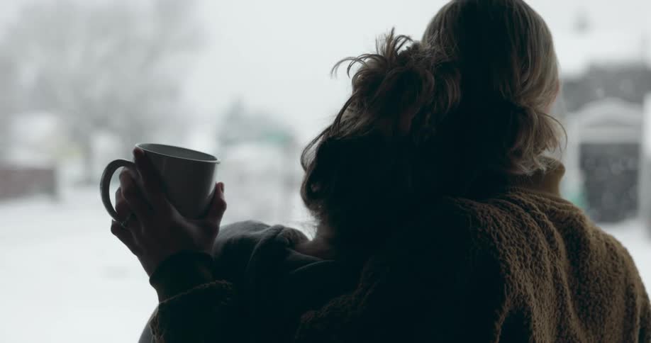 Blonde women holding each other and white cup look out the window while snowing