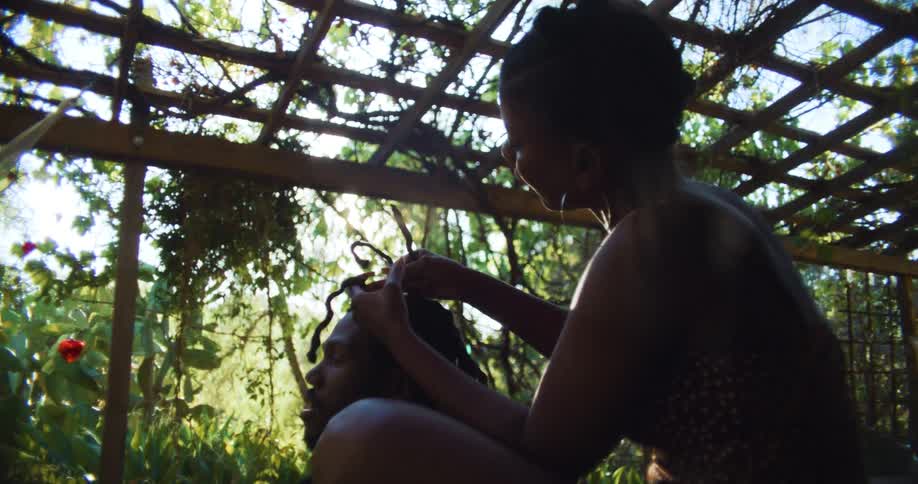 Sunlight shines through plants as woman plaits mans hair as they sit under a lattice work