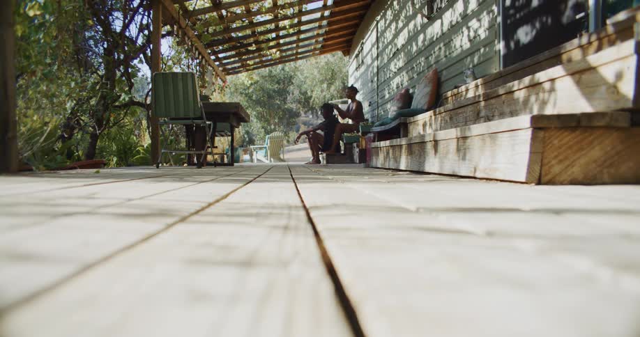 A man and woman sit on a porch and the woman braids his hair