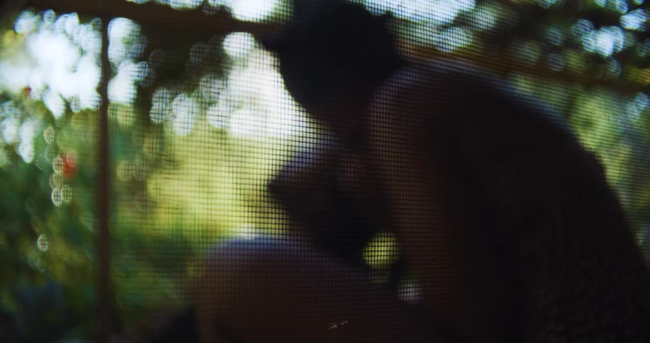 Woman strokes mans head and kisses him as they sit outside under vine covered lattice roof